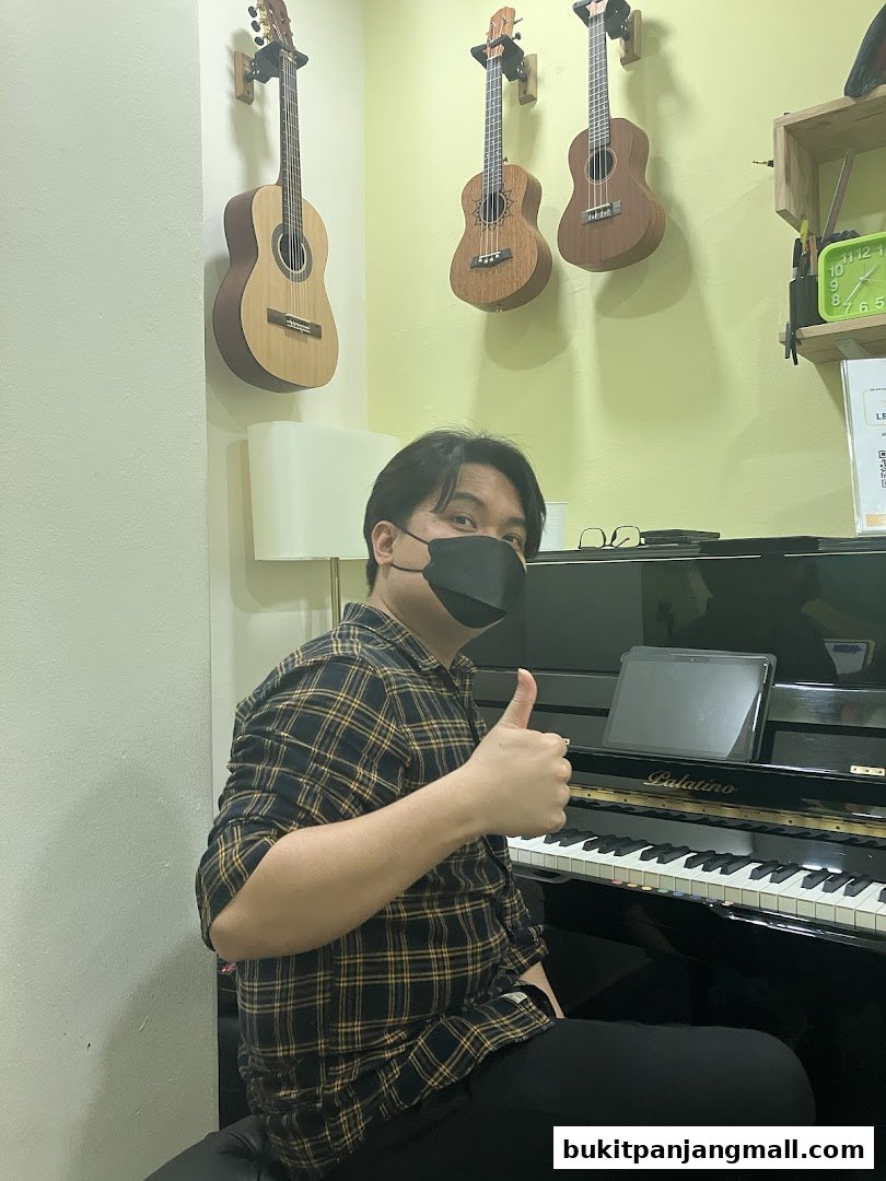 A person wearing a mask gives a thumbs up while sitting at a piano with guitars on the wall.
