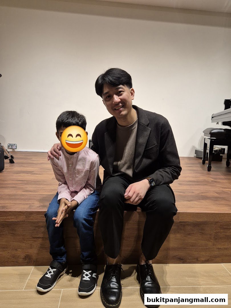 A smiling man and a child sit on a wooden stage with a piano in the background.