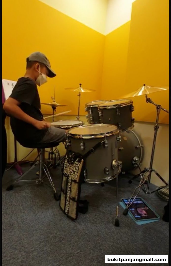 A person practices drumming in a bright yellow room with a drum set and tablet.