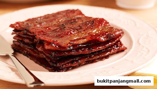 A stack of glistening, sweet bak kwa slices on a pink plate with a serving utensil.