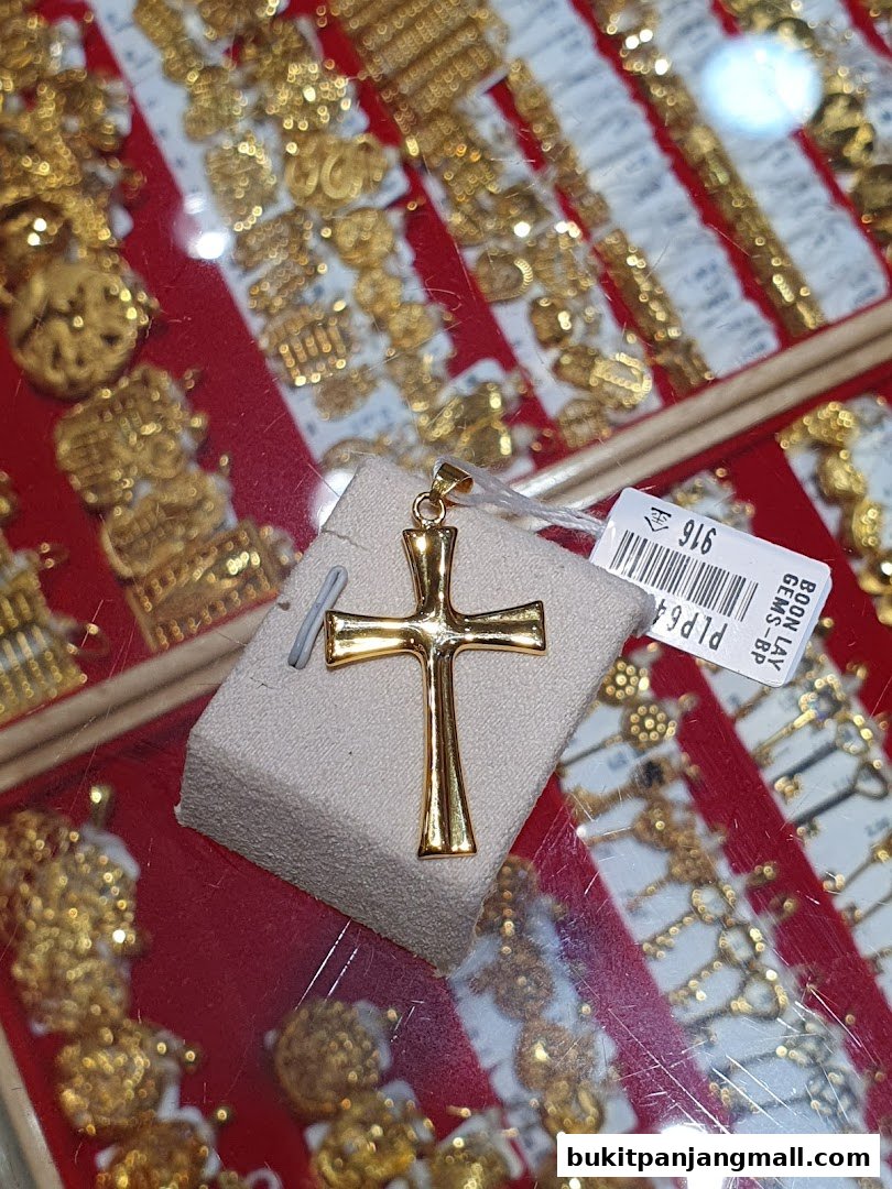 A close-up of a gold cross pendant displayed on a stand, with other gold jewelry in the background.