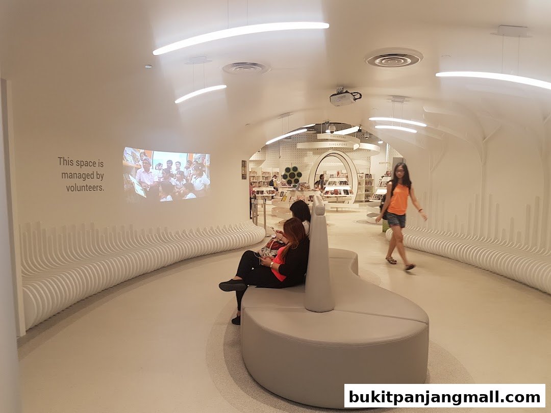 Interior of a modern library with people sitting and walking, featuring curved benches and a projected image.