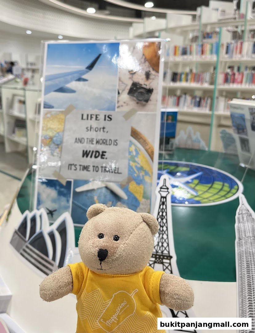 A teddy bear in a yellow shirt stands in front of a display about travel, with bookshelves in the background.