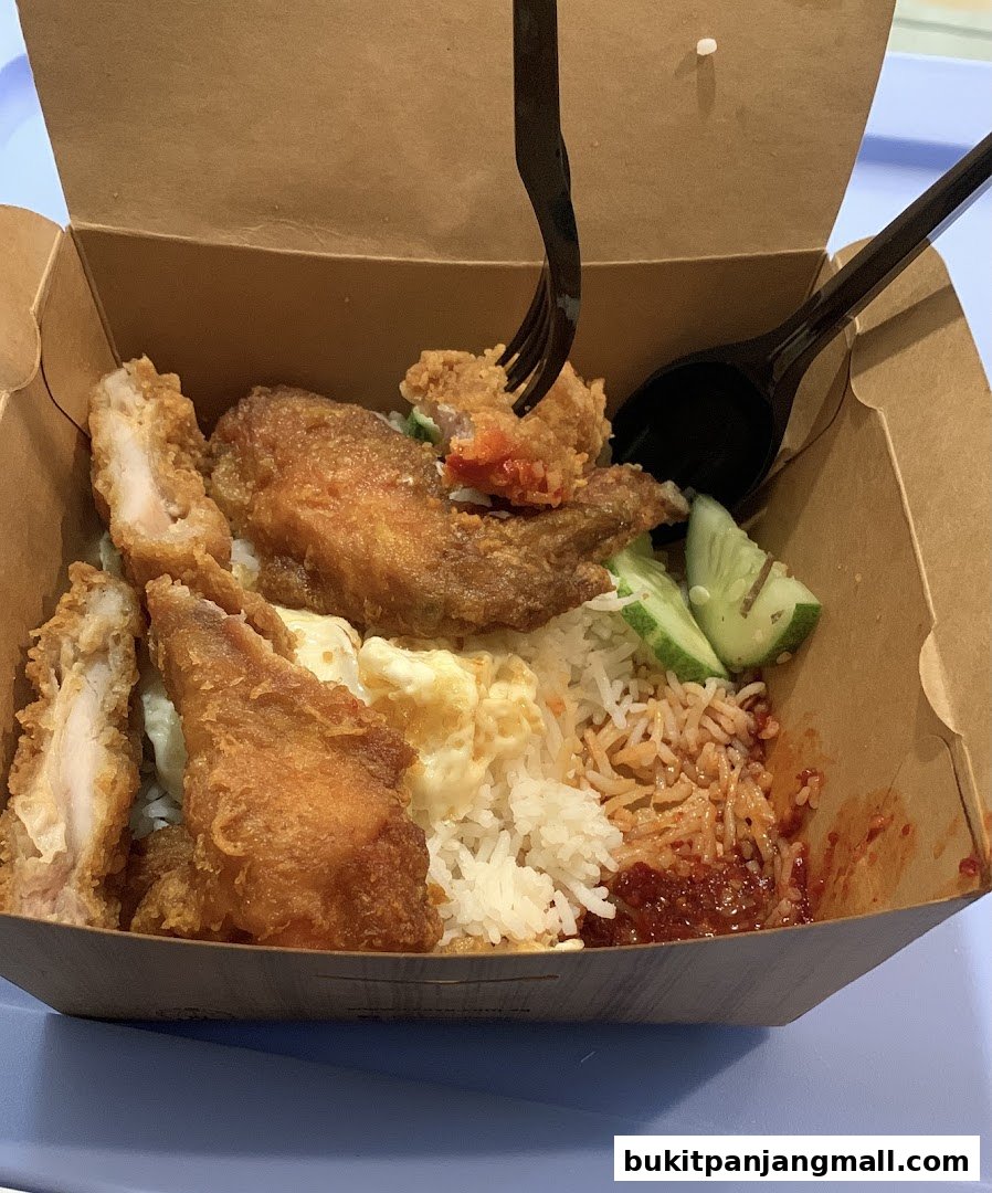 A close-up of a takeaway box filled with Nasi Lemak, featuring fried chicken, rice, and sambal.