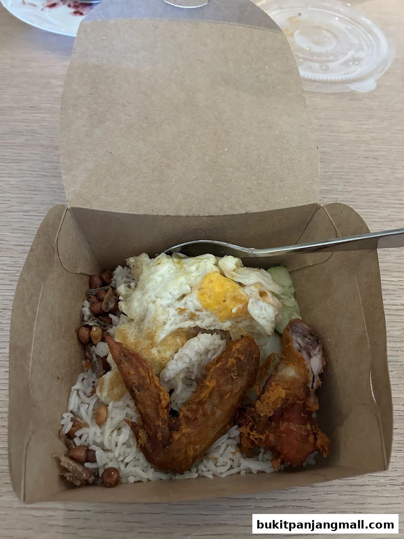 A takeaway box filled with Nasi Lemak, featuring rice, fried chicken, egg, peanuts, and anchovies.