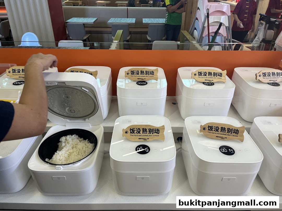 A person's hand opens a rice cooker filled with cooked rice, with several other rice cookers behind it.