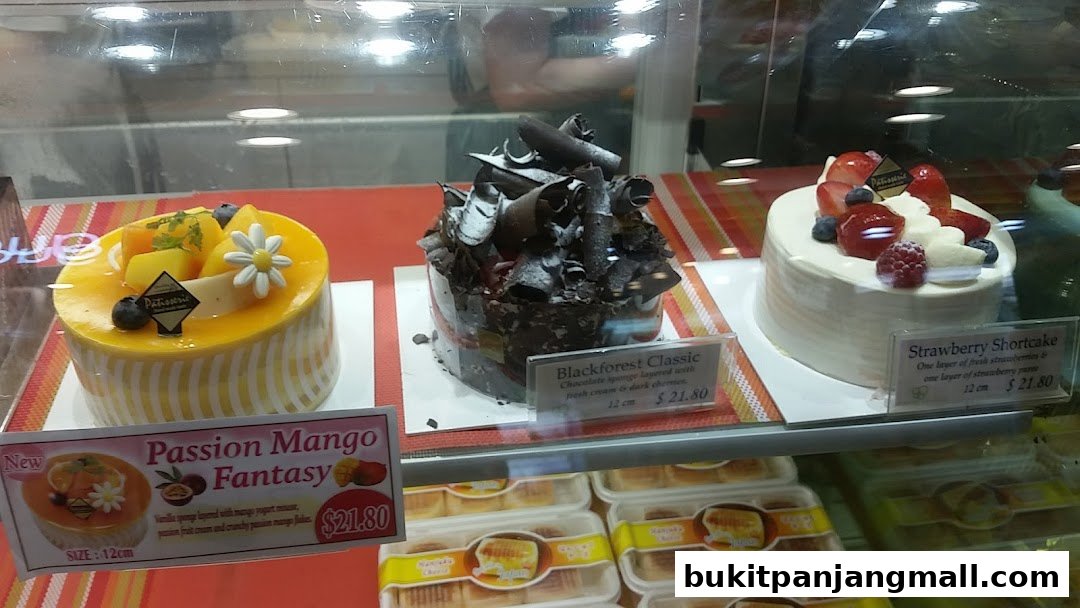 A display case filled with various cakes, including Passion Mango Fantasy, Blackforest Classic, and Strawberry Shortcake.