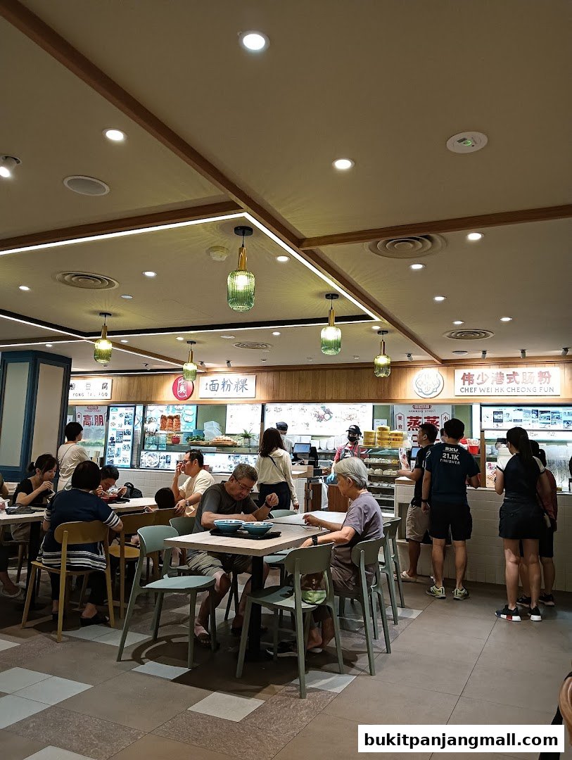 People dining at tables in a busy food court with various food stalls visible.