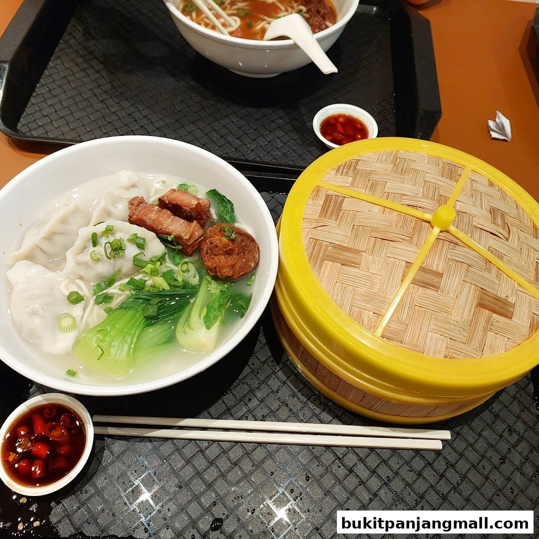 A bowl of wonton noodle soup with meat and vegetables, next to a bamboo steamer.