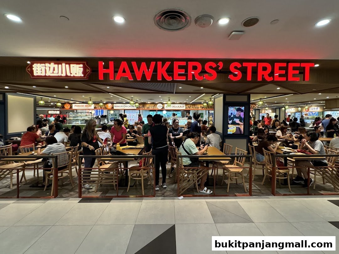 A busy hawker street food court with many people dining at tables under a large illuminated sign.