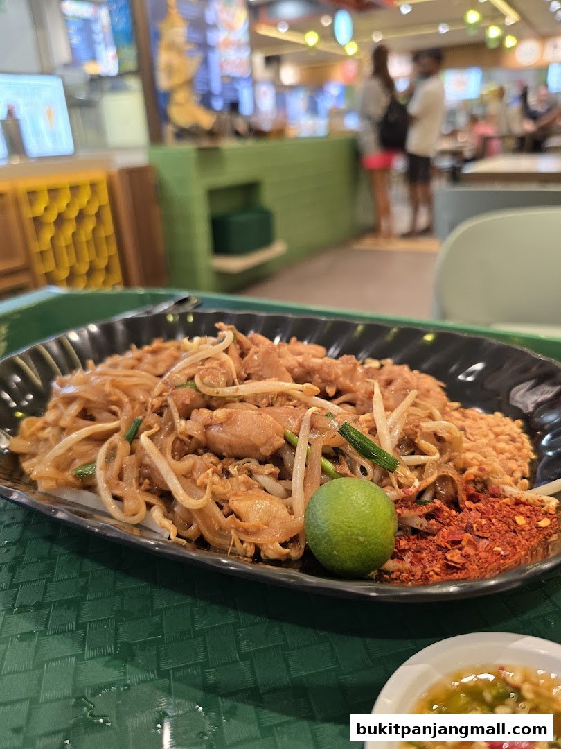 A plate of Pad Thai with lime and chili flakes, served in a food court setting.
