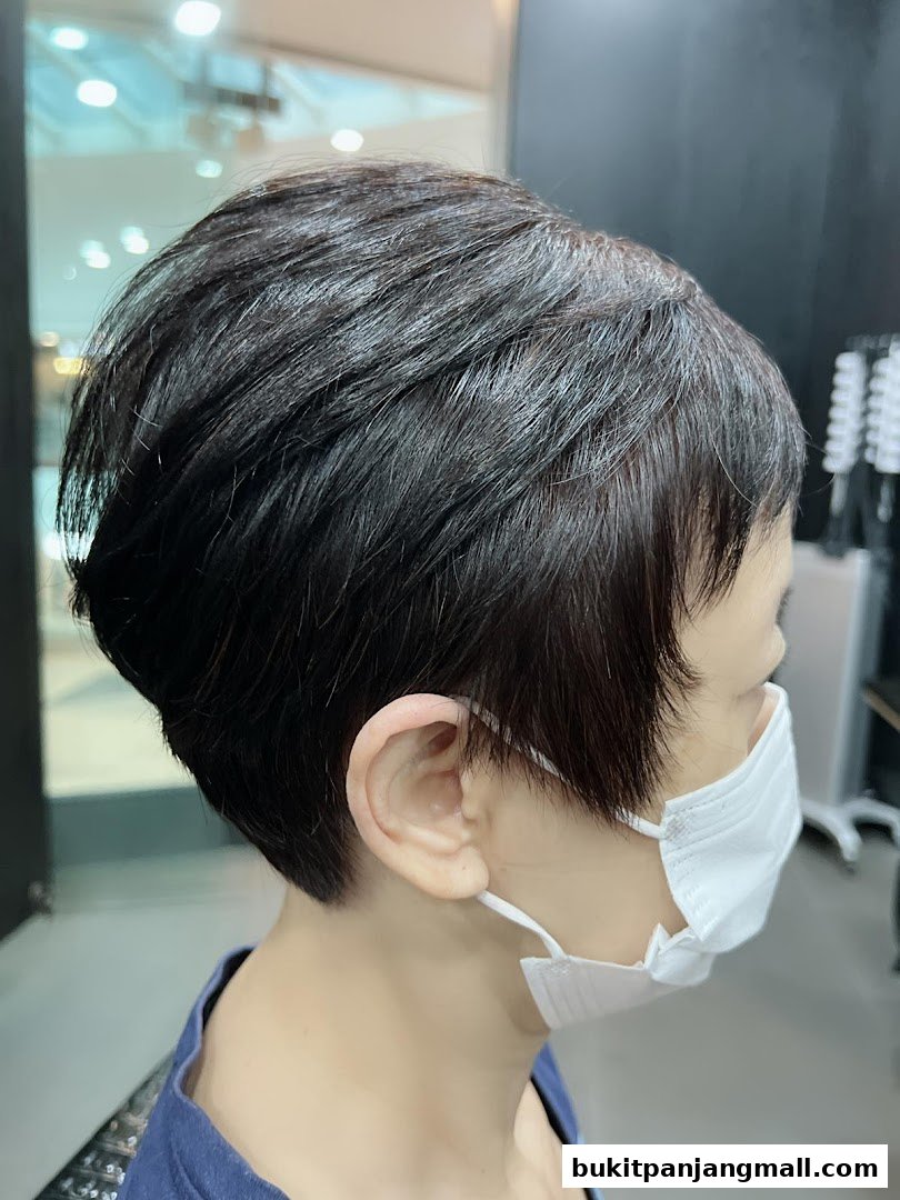 A close-up profile view of a person with a short, dark haircut wearing a white face mask.