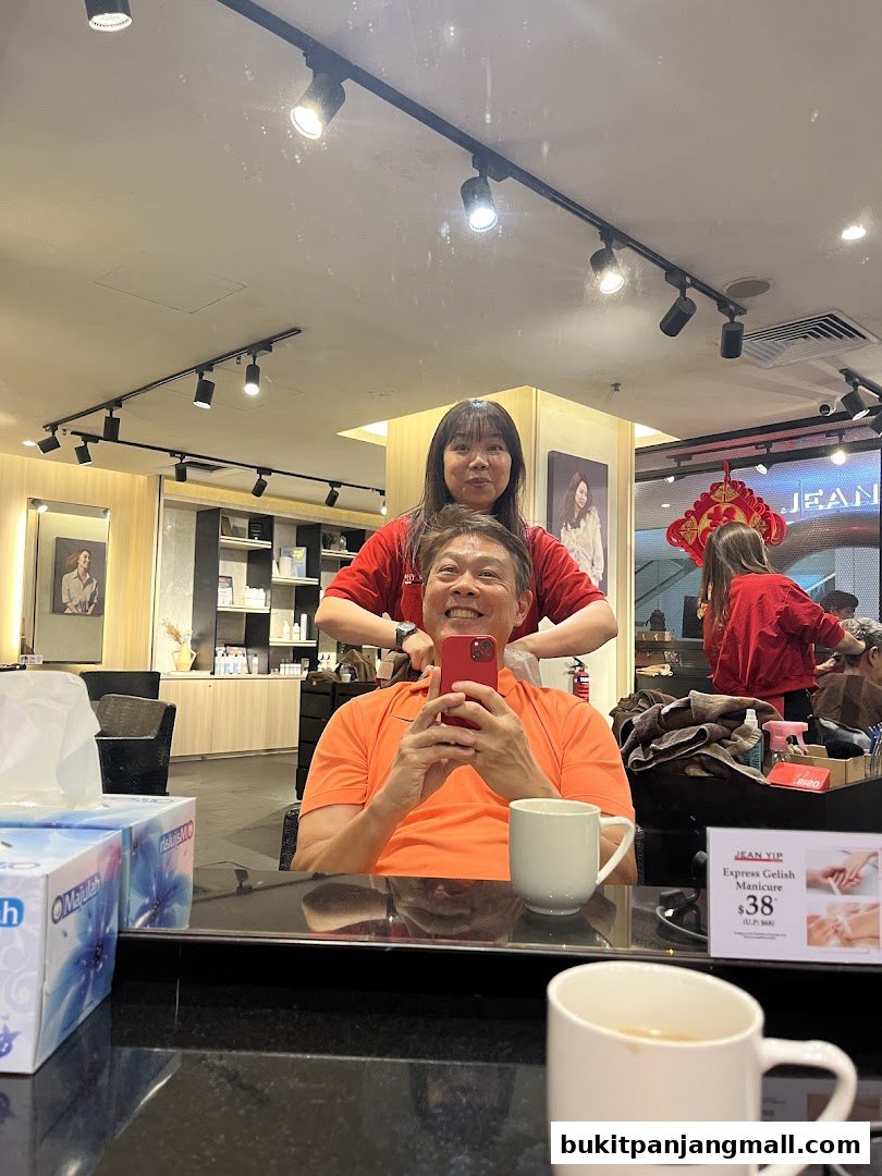 A man smiles while taking a selfie in a salon as a stylist works on his hair.