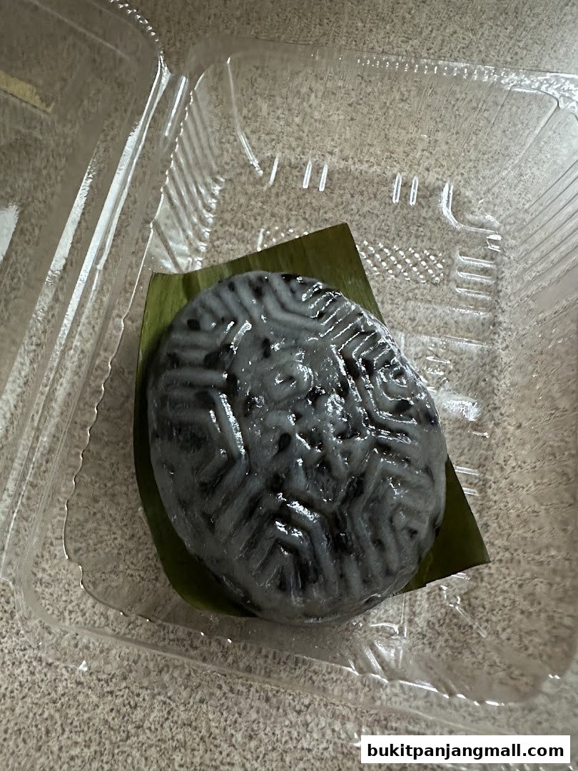 A close-up of a black Ang Ku Kueh with a patterned top, served in a clear plastic container.