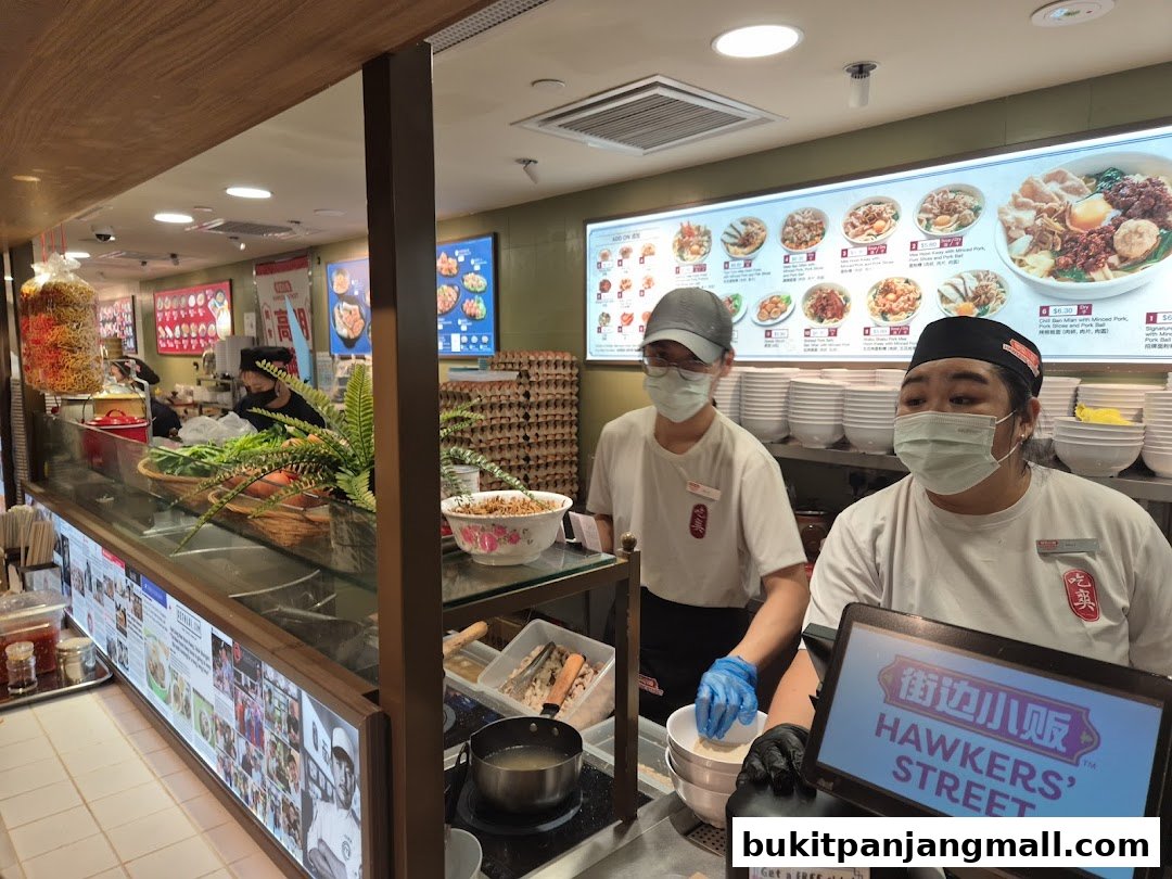 Two staff members wearing masks stand behind a counter at a food stall, with a menu board visible.