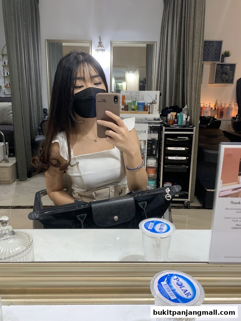 A woman takes a selfie in a salon mirror, showing hair products and styling equipment.