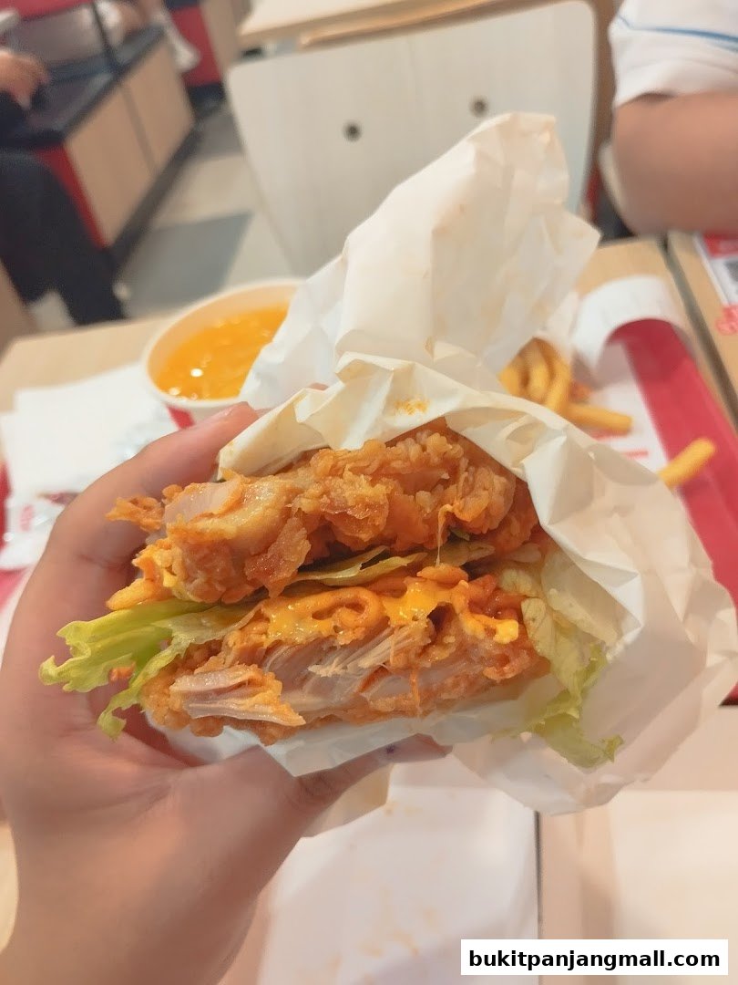 A person holds a juicy KFC burger with melted cheese and lettuce, with fries and a drink in the background.