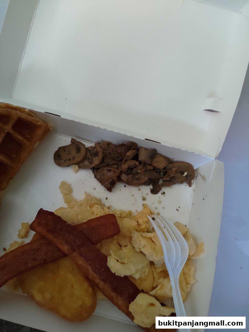 A takeaway box filled with a breakfast meal including waffles, scrambled eggs, hash browns, and mushrooms.