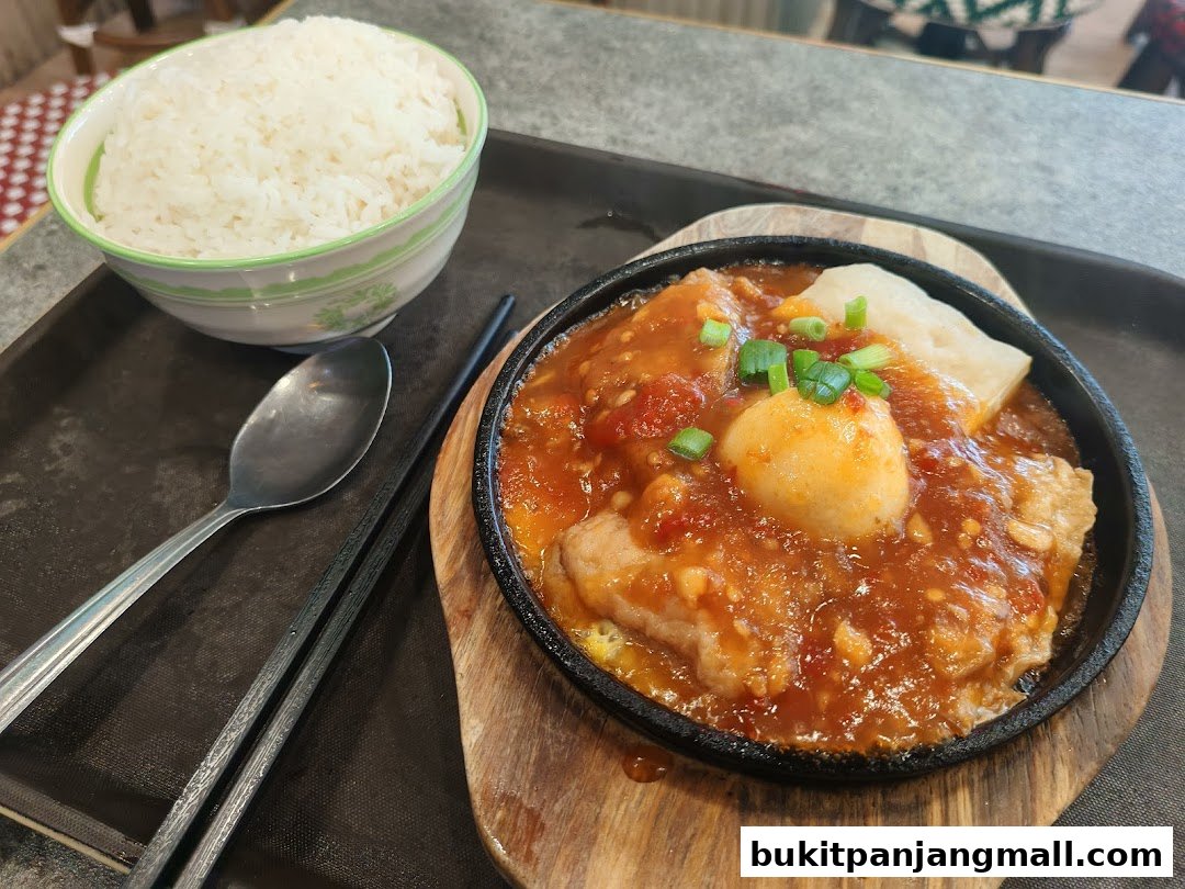A sizzling dish with sauce, tofu, and a meatball, served with a bowl of rice.
