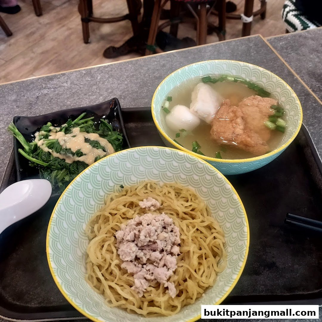 A close-up of a meal at Koo Kee, featuring noodles, soup with fish cakes, and a side of greens.