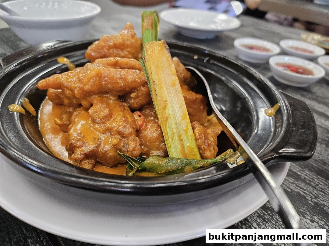 A close-up of a dish with crispy fried pieces coated in a rich, orange sauce, garnished with lemongrass.