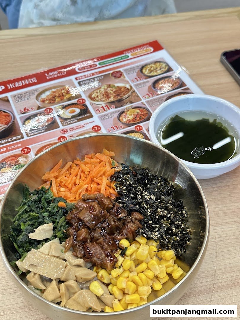 A close-up of a bibimbap dish with a menu and soup in the background.