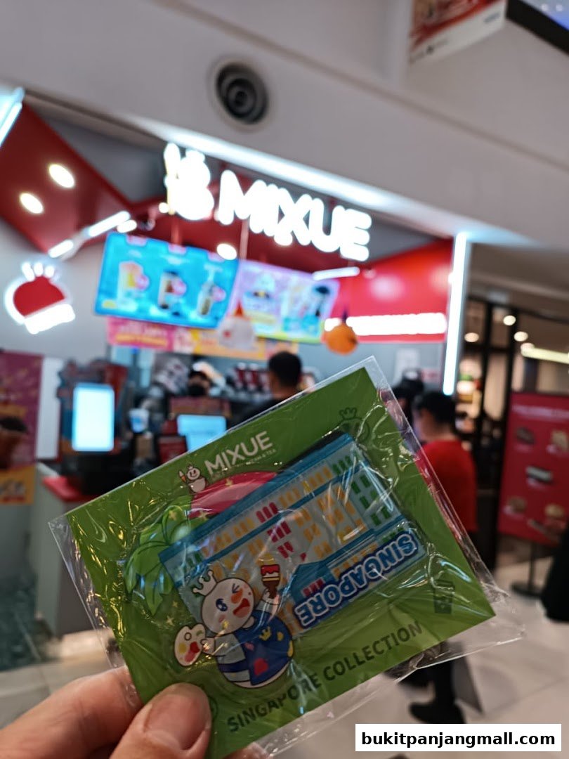A hand holds a souvenir magnet in front of a brightly lit MIXUE ice cream and tea shop.