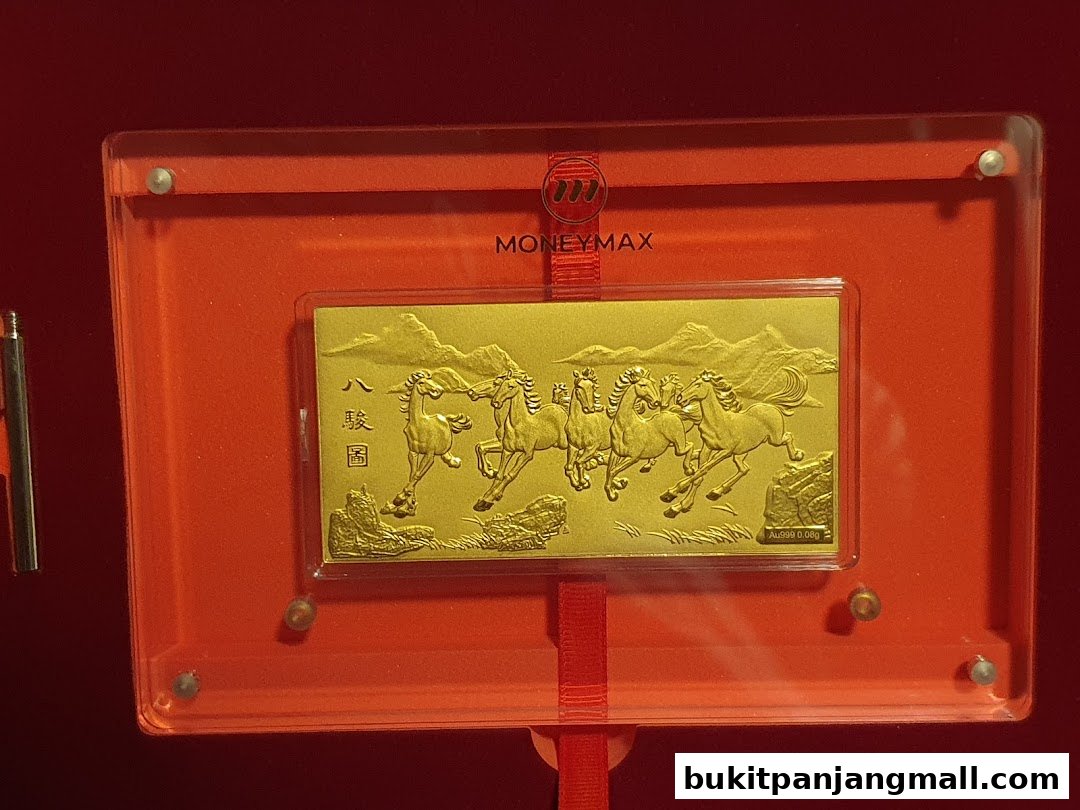 A gold bar with embossed horses displayed in a red frame with the MoneyMax logo.