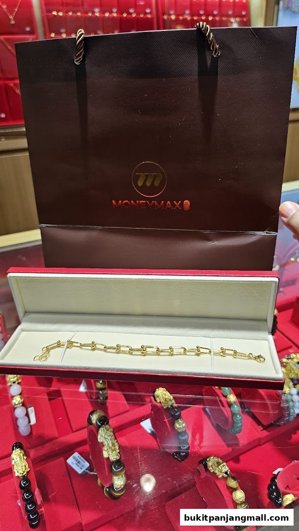 A gold bracelet displayed in a red box with a MoneyMax shopping bag in the background.