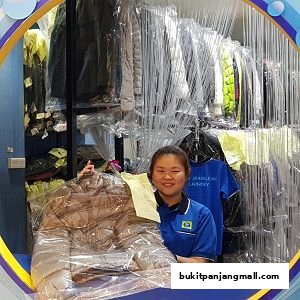A smiling staff member stands behind a counter at a laundry shop, holding a garment.