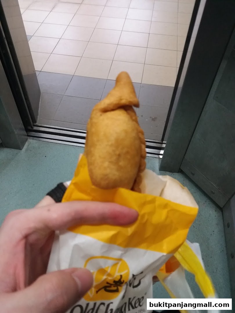A hand holds a freshly fried curry puff from Old Chang Kee in a branded paper bag.