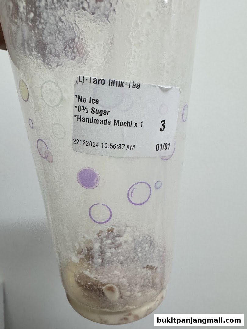 A close-up of a plastic cup with a label indicating a Taro Milk Tea with no ice and 0% sugar.