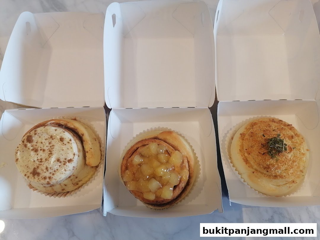 Three freshly baked cinnamon rolls in white boxes, one with cream cheese frosting, one with apple filling, and one with a savory topping.