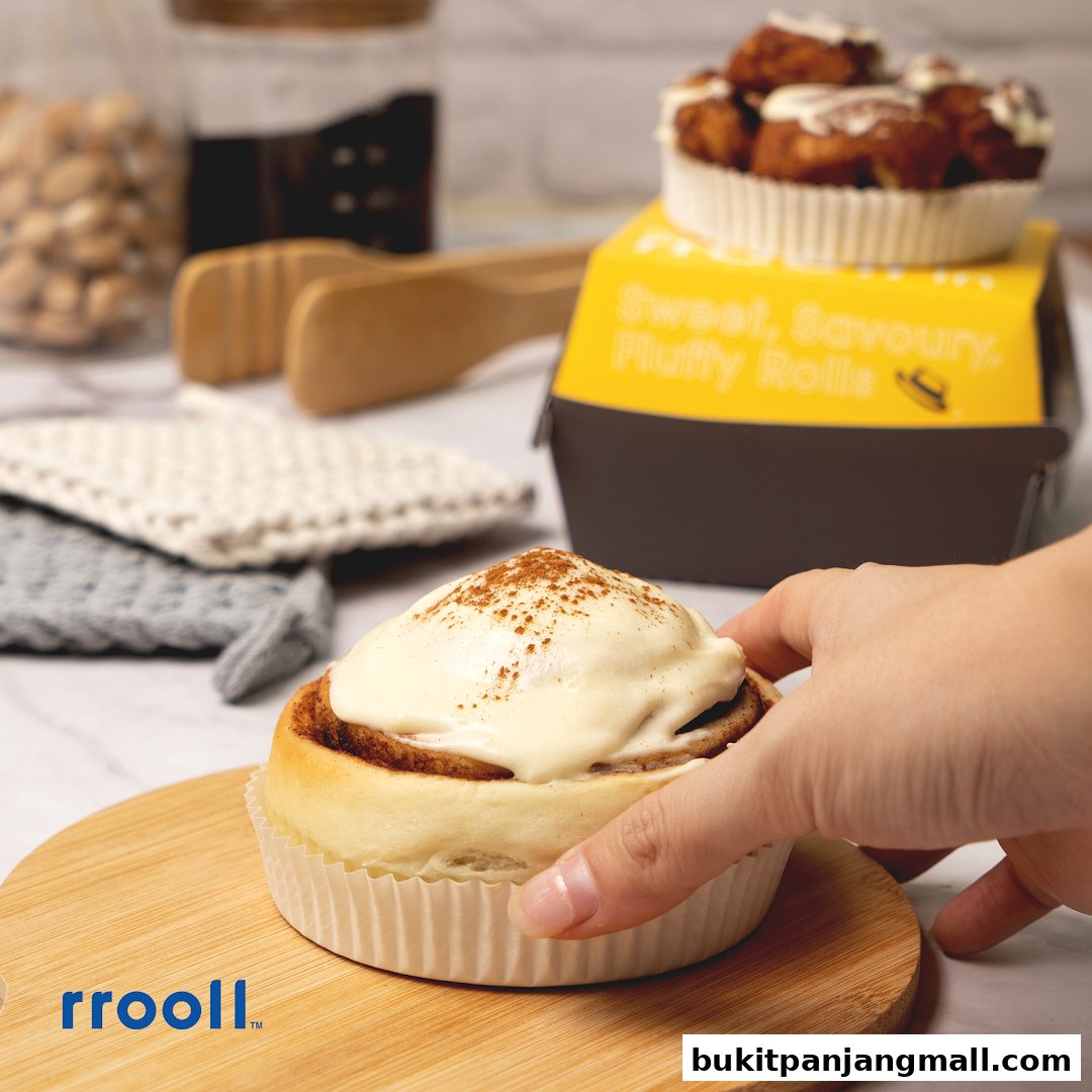 A hand reaching for a frosted cinnamon roll with a box of rolls in the background.