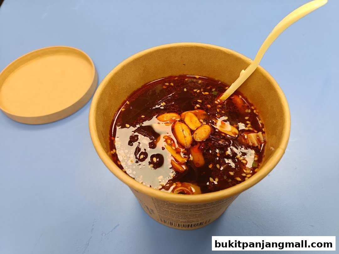 A cup of spicy noodle soup with peanuts and sesame seeds, served with a plastic spoon.