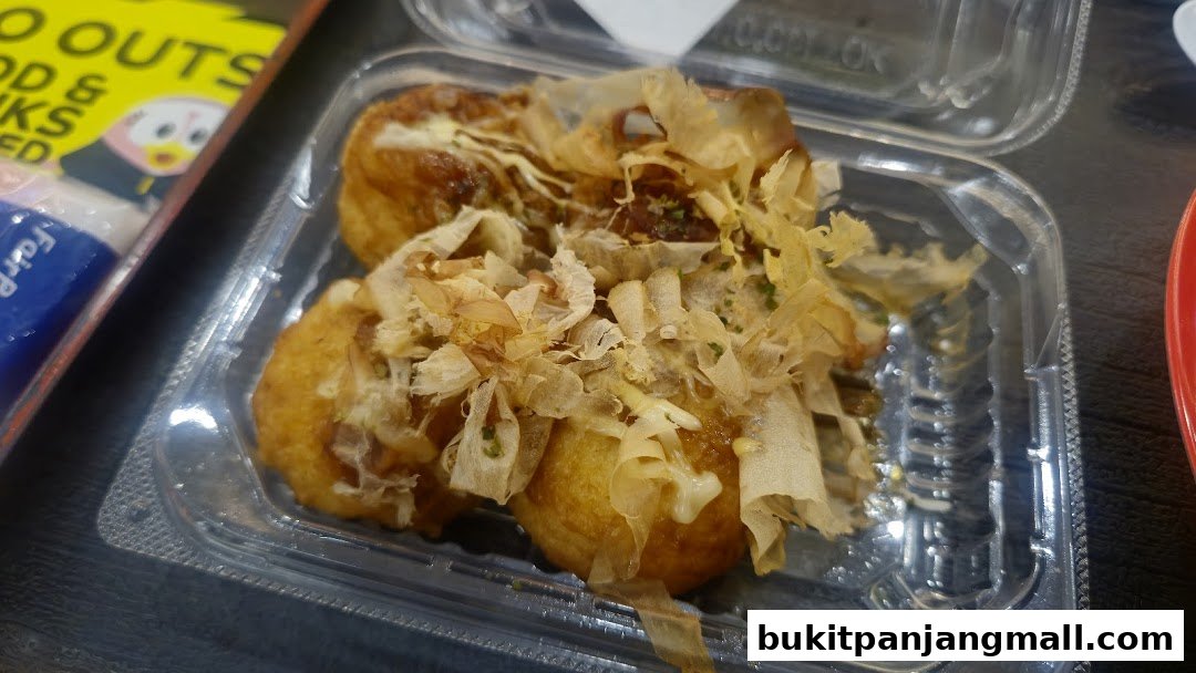 A close-up of takoyaki in a clear plastic container, topped with bonito flakes.