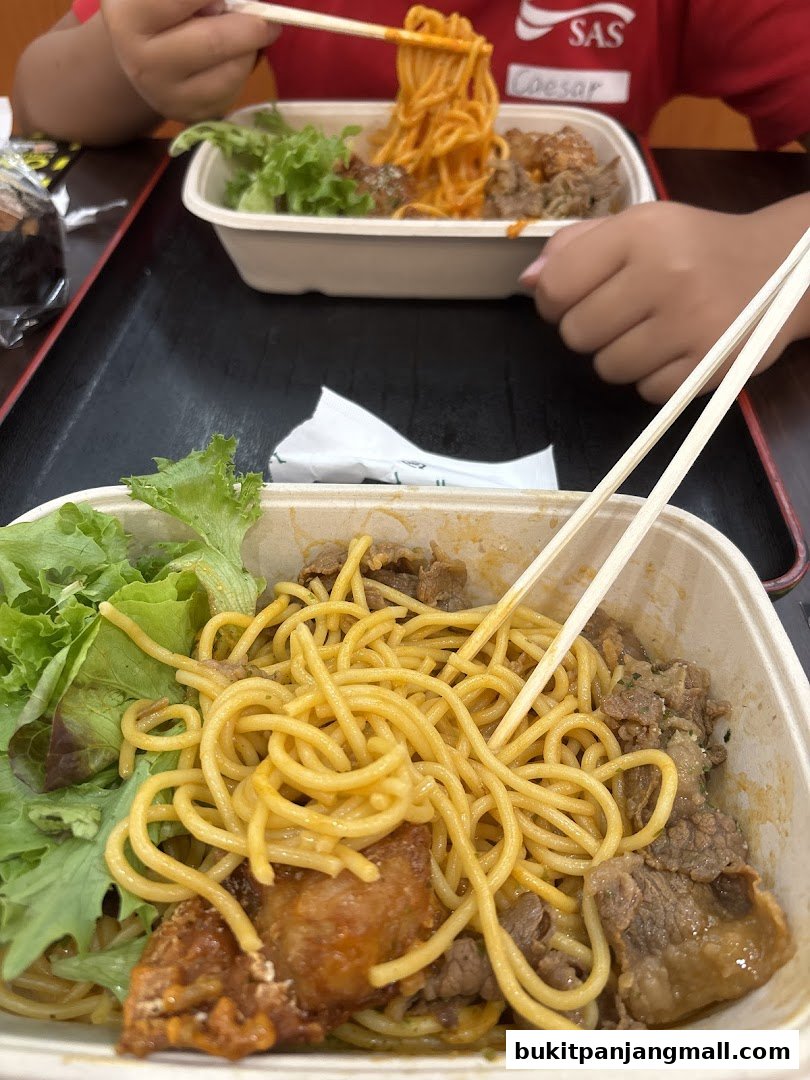 Close-up of two takeaway pasta dishes with meat and salad, being eaten with chopsticks.