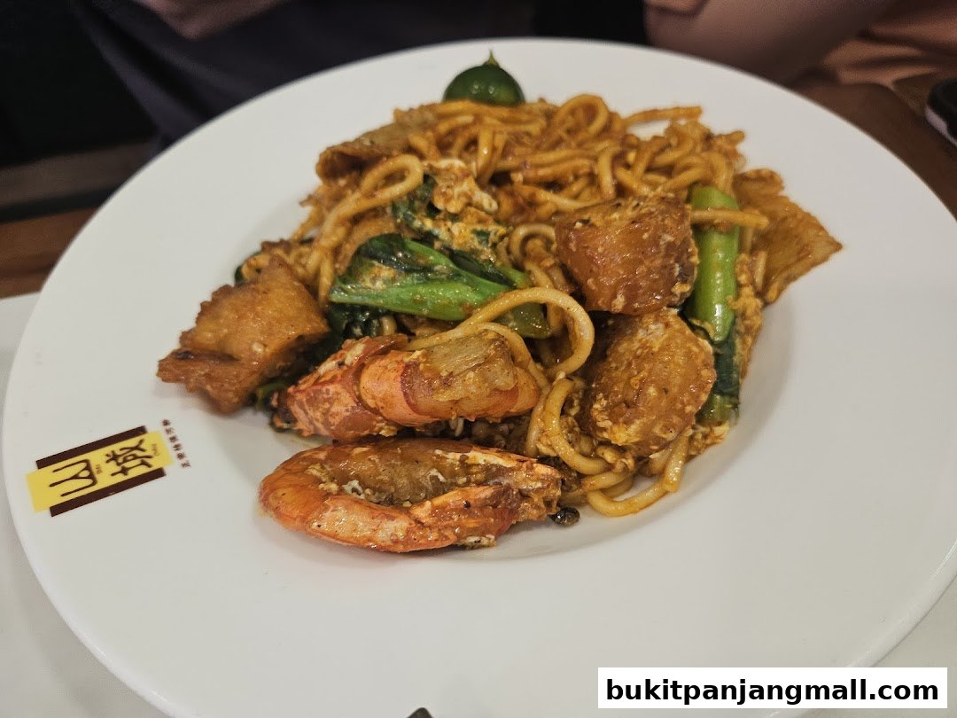 A plate of stir-fried noodles with prawns, fried meat, and vegetables, garnished with a lime wedge.