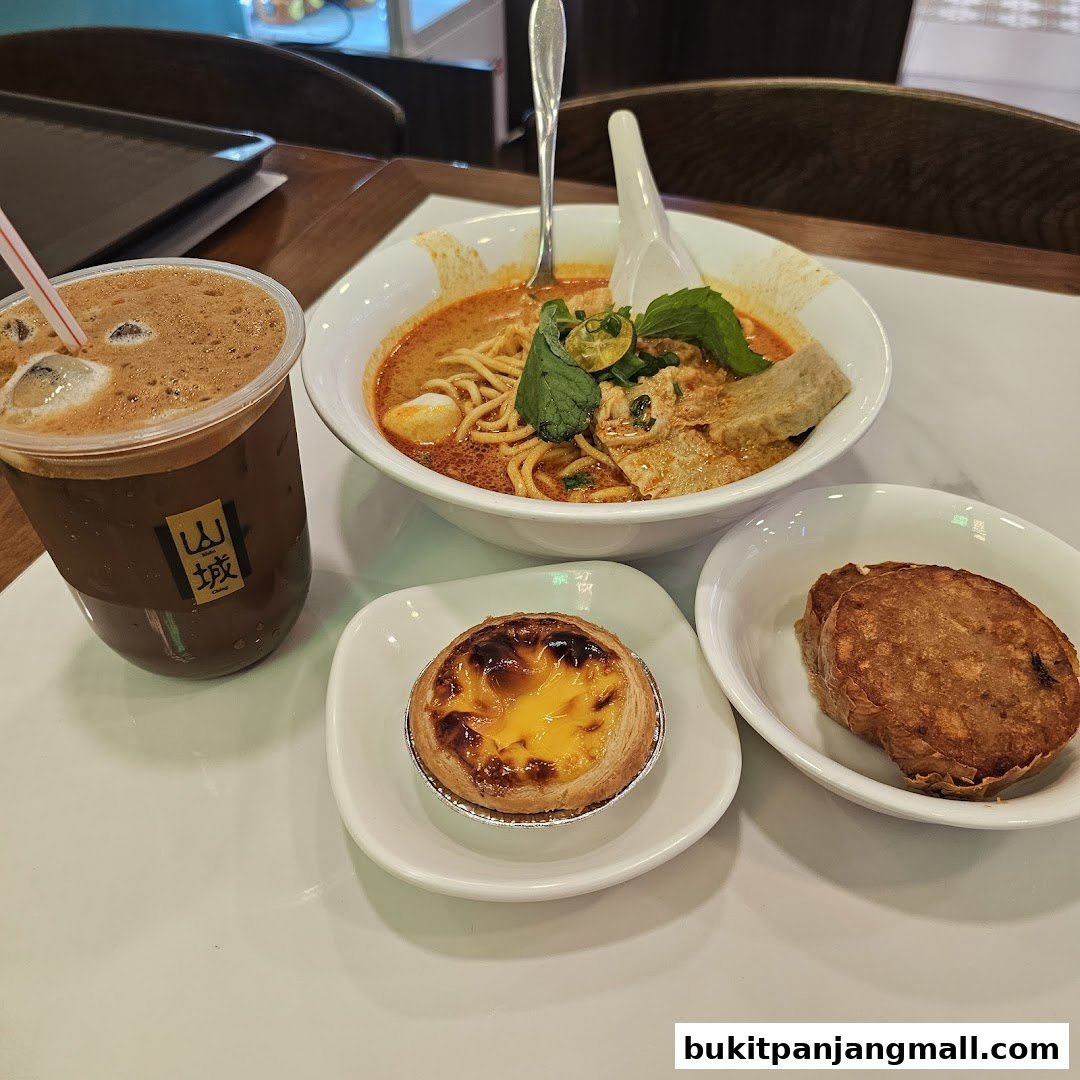A bowl of laksa, an egg tart, a fried item, and a drink are displayed on a white table.