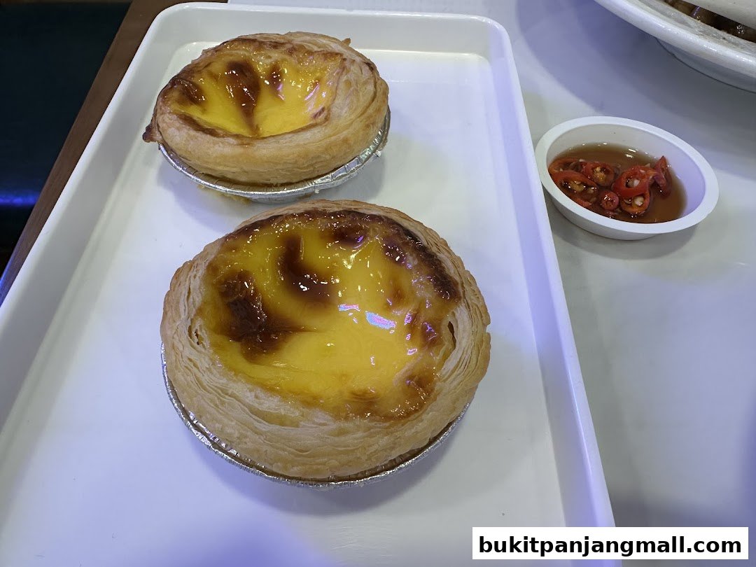 Two golden-brown egg tarts with a creamy custard filling, served with a side of chili sauce.