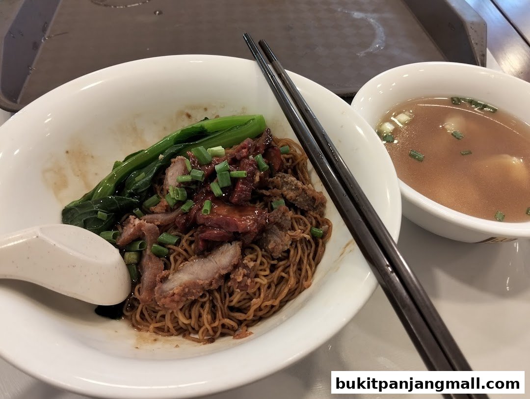 A bowl of Ipoh style noodles with char siu, vegetables, and a side of soup.