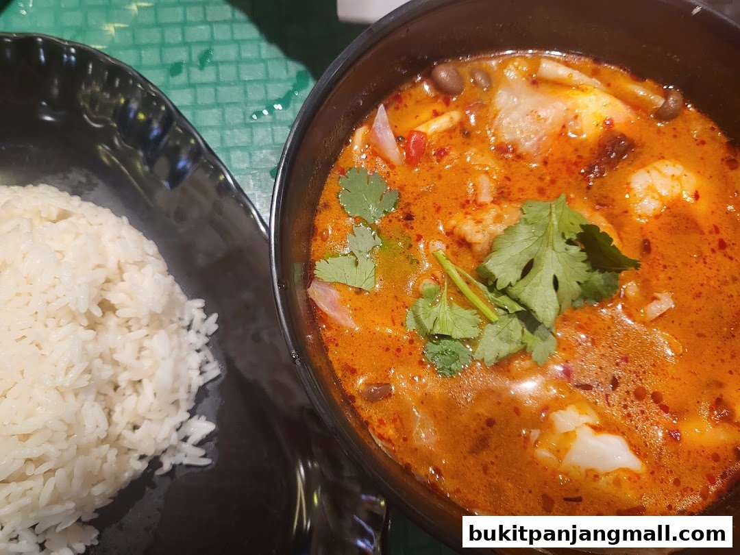 A close-up of a steaming bowl of Tom Yum soup with rice, garnished with cilantro.