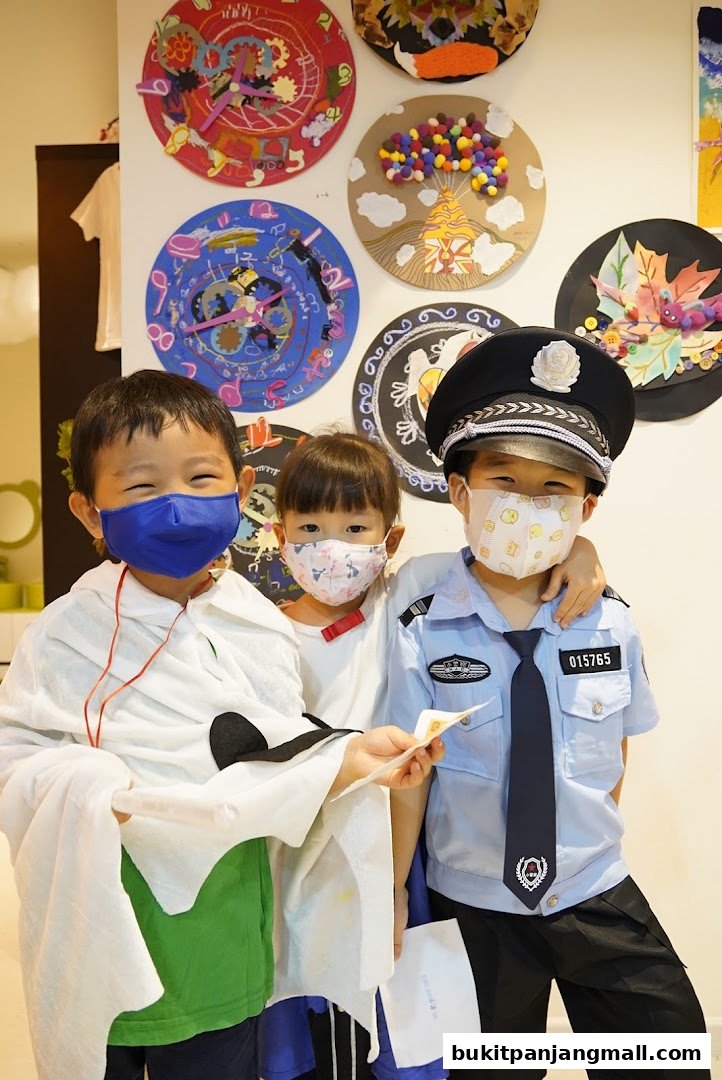 Three children in costumes, one as a police officer, pose in front of colorful artwork.