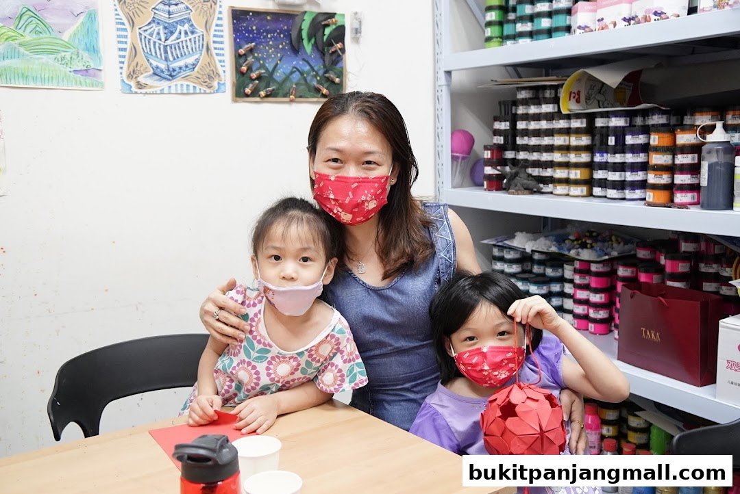 A woman and two children wearing masks pose in an art studio with shelves of art supplies.