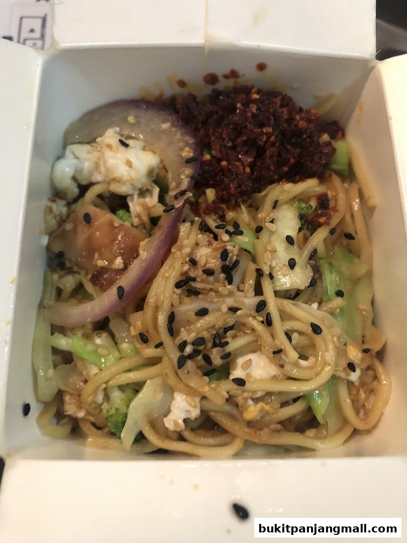 A close-up of a takeaway box filled with noodles, vegetables, and chili flakes.
